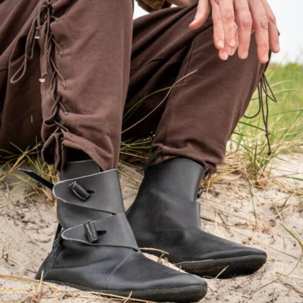 Black leather Viking boots with buckle straps, worn with brown pants on a sandy ground.
