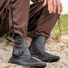 Black leather Viking boots with buckle straps, worn with brown pants on a sandy ground.