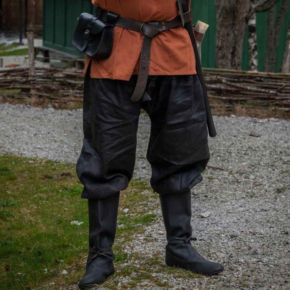 Man wearing black linen Viking pants tucked into tall leather boots, paired with a leather belt and pouch outdoors.