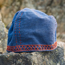 Blue cotton Viking cap with red embroidered Norse motifs, resting on a rock.