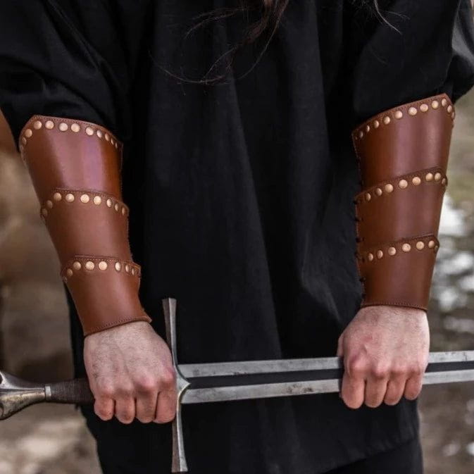 Brown leather Viking arm bracers with rugged rivets and adjustable straps, worn by a person holding a sword.