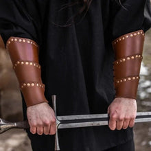 Brown leather Viking arm bracers with rugged rivets and adjustable straps, worn by a person holding a sword.