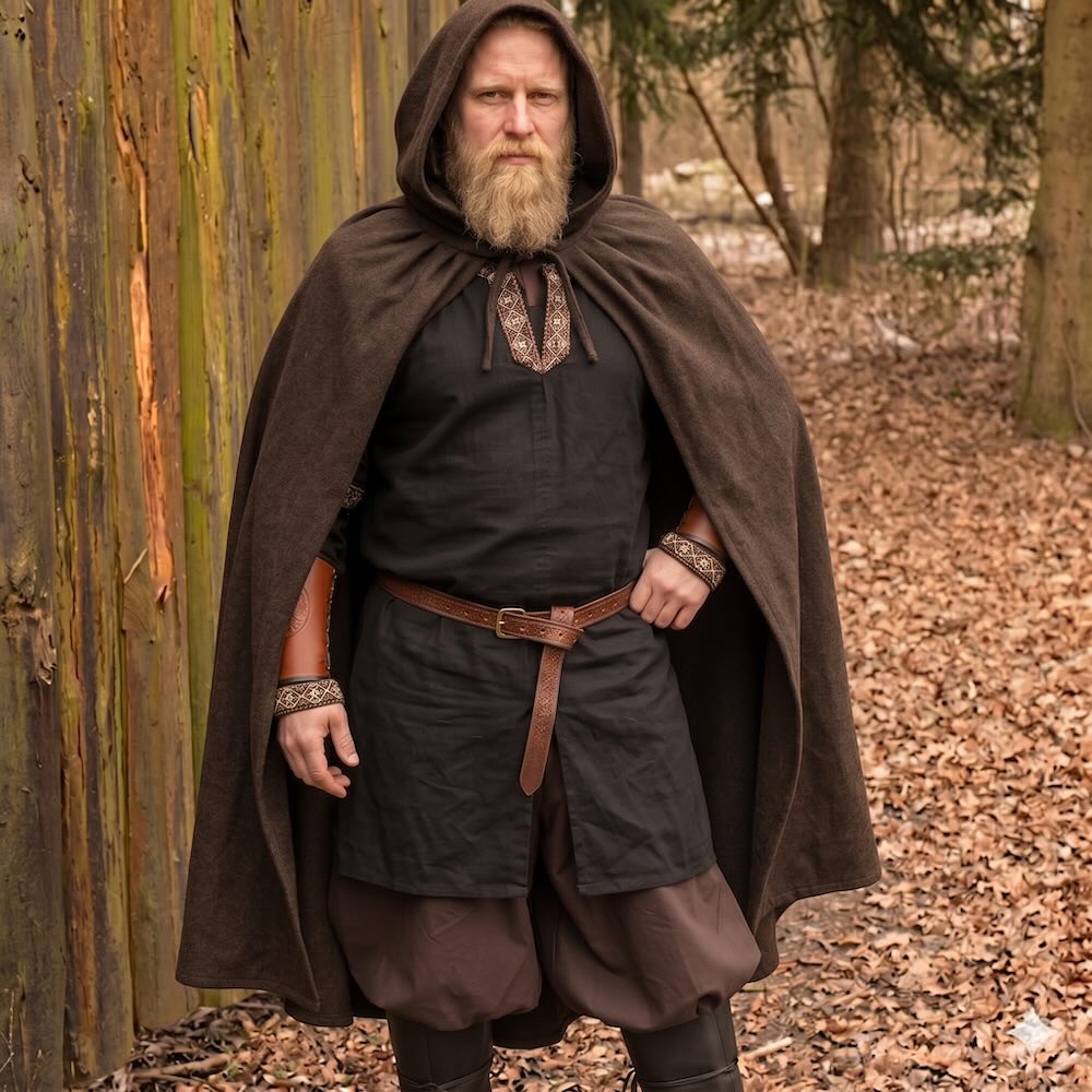 Viking warrior in a brown hooded wool cloak, black tunic with embroidered trim, leather belt, and bracers.