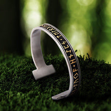 Viking Elder Futhark rune bracelet, silver-toned metal with black and gold engraved runes, on moss.