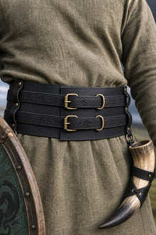 A close-up of a Viking wearing a wide black leather Viking-style belt with two brass buckle details, over an olive green linen tunic. A horn is tucked into the belt loop on the right side.