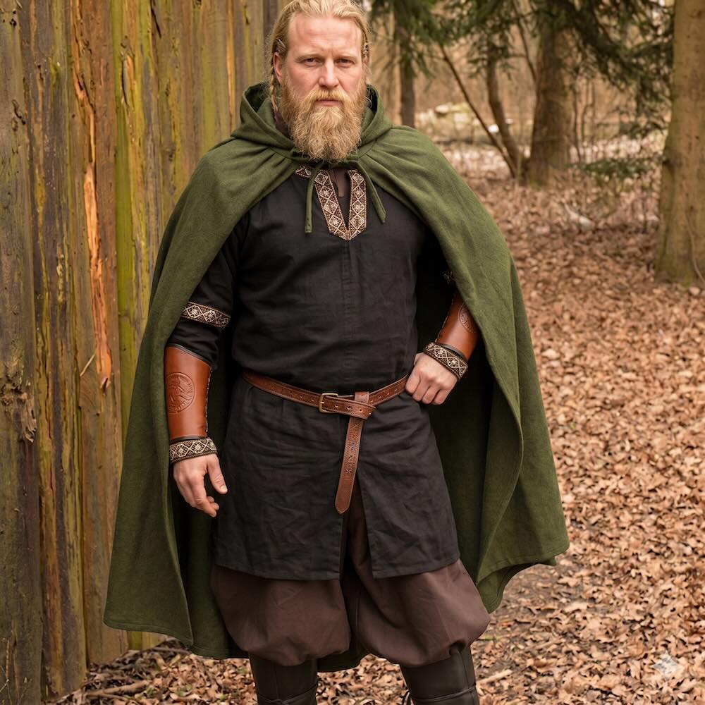Viking warrior in a hooded, dark green wool cloak over a black tunic with embroidered trim and brown leather bracers.
