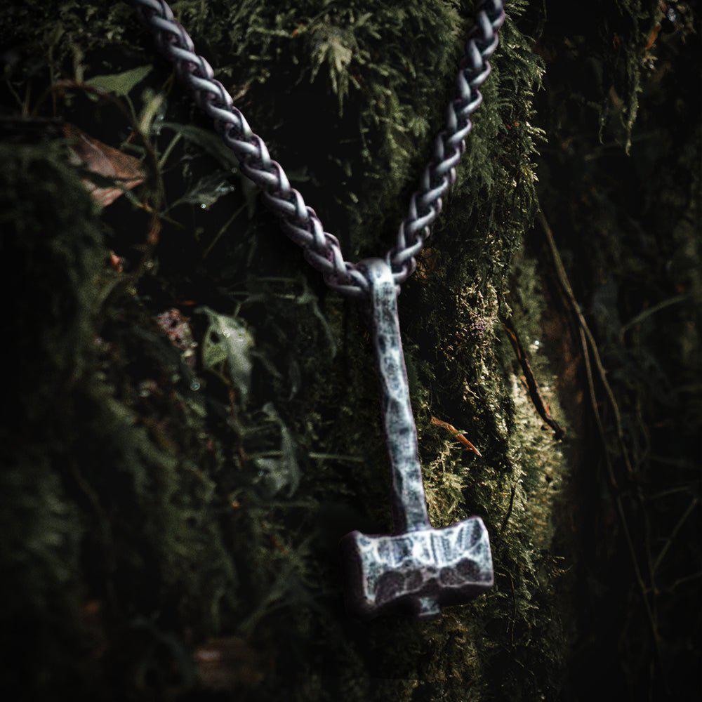 Viking hammered iron Thor's Hammer Mjolnir pendant on a thick chain, against mossy bark.