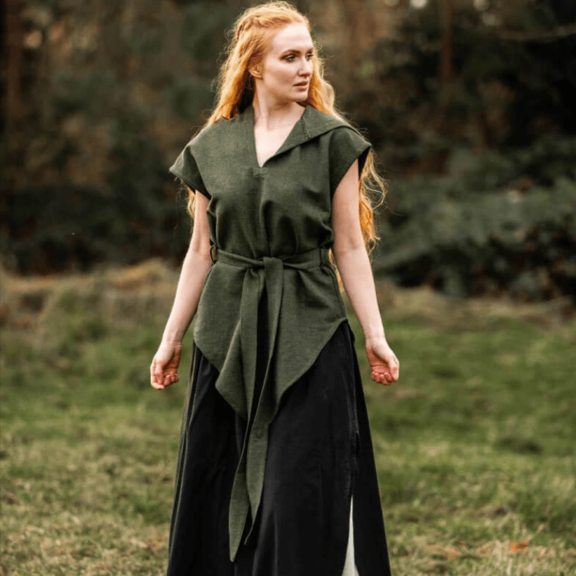 Woman in a hooded green Viking tunic with a belt, over a black skirt, in a field.