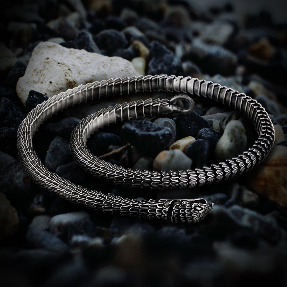 Viking Jormungandr segmented steel necklace, silver-toned, coiled on dark pebbles.