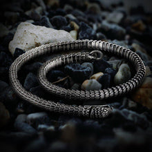 Viking Jormungandr segmented steel necklace, silver-toned, coiled on dark pebbles.