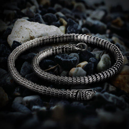 Viking Jormungandr segmented steel necklace, silver-toned, coiled on dark pebbles.