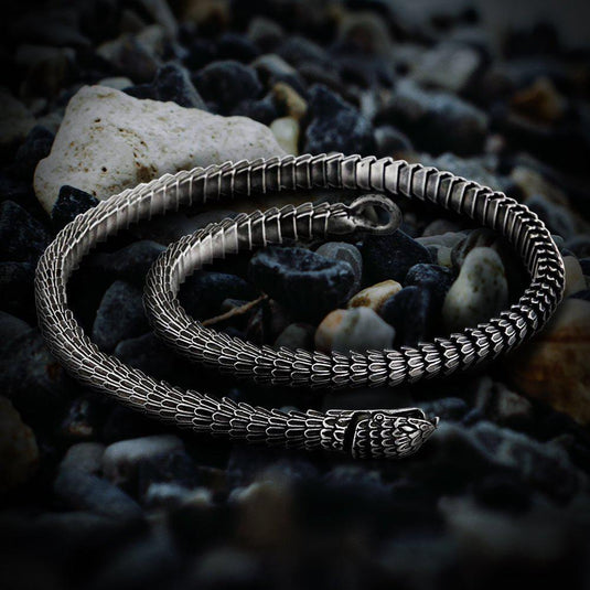 Viking Jormungandr segmented steel necklace, silver-toned, coiled on dark pebbles.