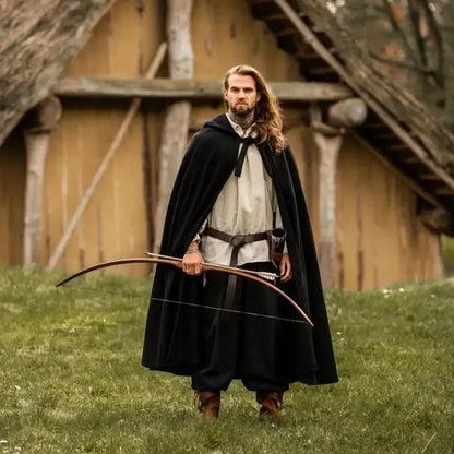 Man in a black hooded Viking wool blend cloak, holding a bow, standing in front of a wooden house.