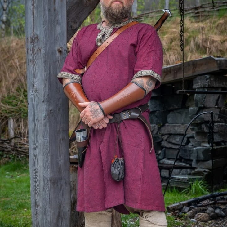 Viking man in a short-sleeved red tunic with embroidered trim, leather bracers, and belt.