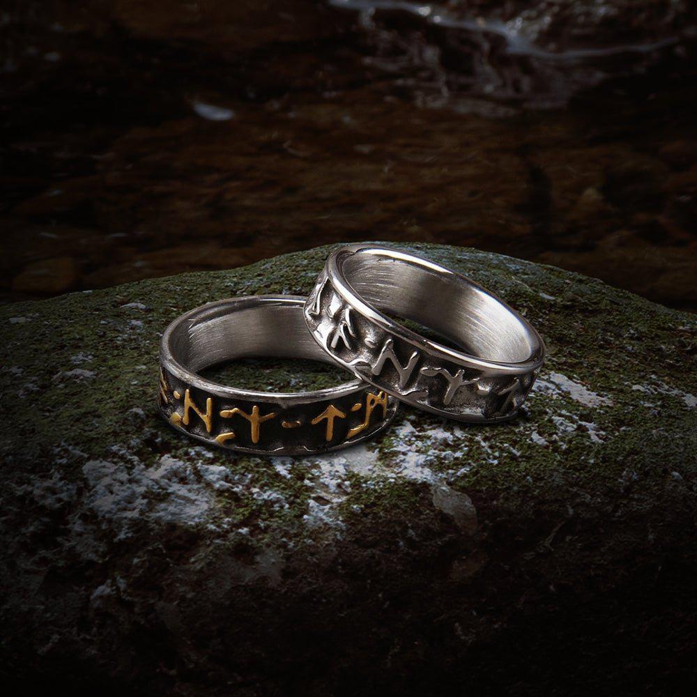 Two bold steel Viking Futhark rune rings on a mossy rock; one with gold runes, one with silver.