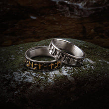 Two bold steel Viking Futhark rune rings on a mossy rock; one with gold runes, one with silver.