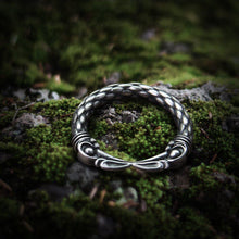 Viking steel ring with Hugin and Munin raven heads, intricate silver knotwork, on mossy ground.