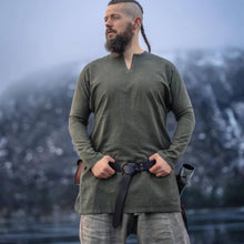 Bearded man wearing olive green linen Viking tunic, leather belt, horn, and pouch, evoking Norse warrior style.