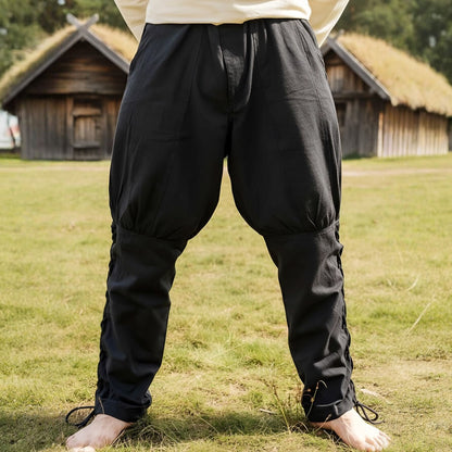 Person wearing Premium Black Viking Pants, Authentic Cut with Leg Lacing, standing on grass with wooden huts in the background