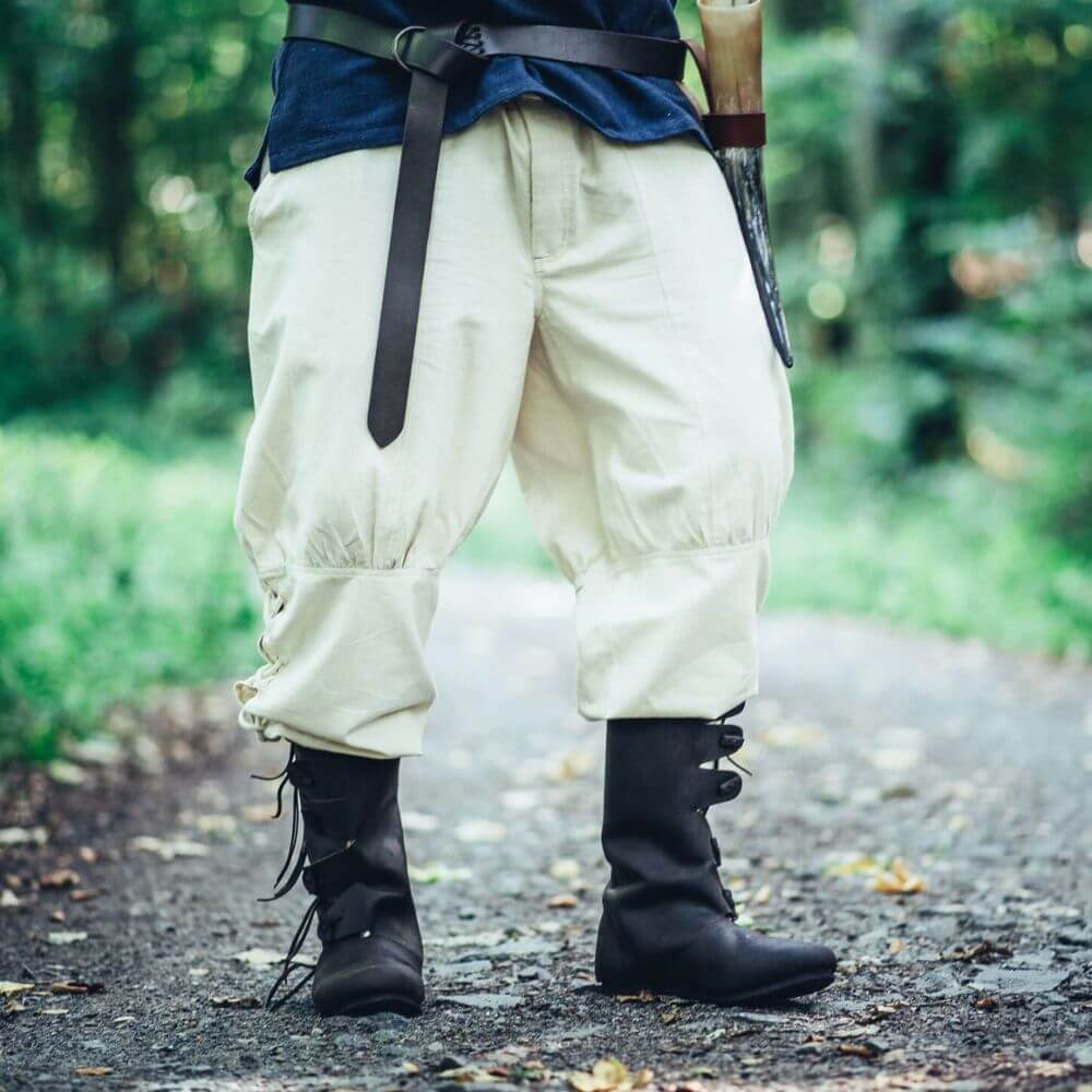 Man standing on a forest path wearing Premium Viking Pants in Natural with tall black boots and a leather belt.