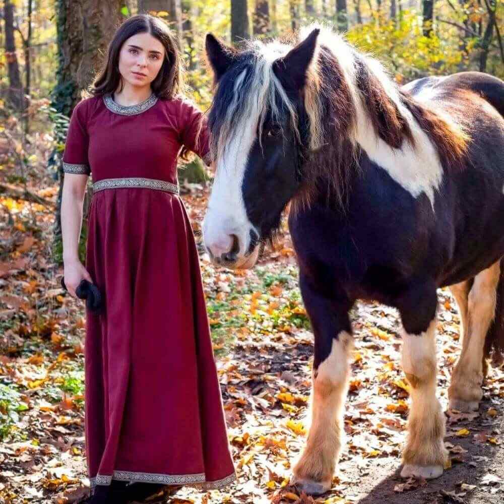 Viking woman in a red short-sleeve dress with embroidered trim, standing next to a horse in a forest.