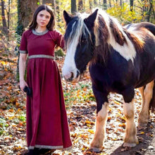 Viking woman in a red short-sleeve dress with embroidered trim, standing next to a horse in a forest.