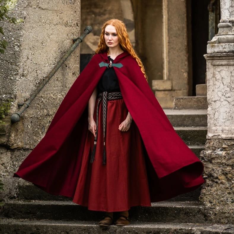 Viking woman in a flowing red wool cloak with a wolf clasp, standing on stone steps.