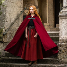 Viking woman in a flowing red wool cloak with a wolf clasp, standing on stone steps.