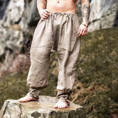 Barefoot man stands on a rock wearing loose, stone color linen Viking pants with drawstring waist and ankle ties.