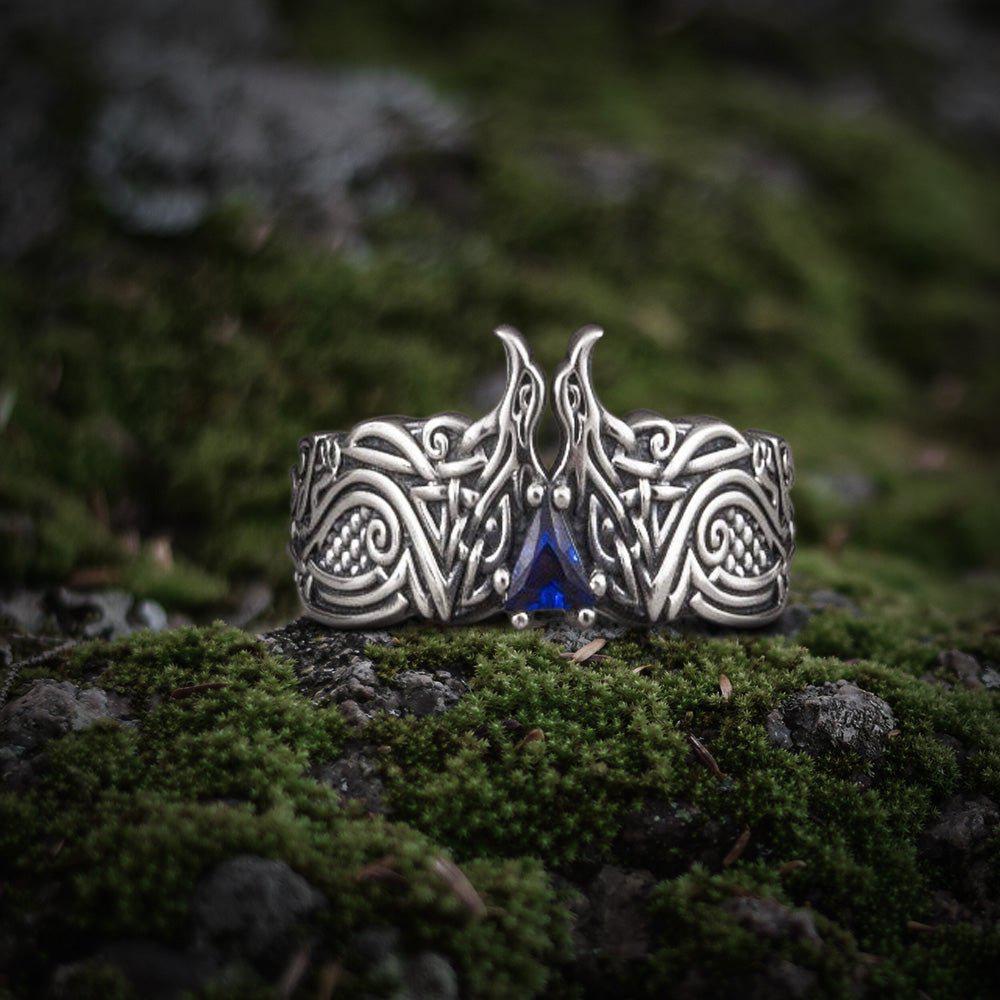 Silver Viking ring with intricate raven design and a triangular blue gem on moss.