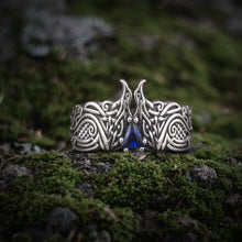 Silver Viking ring with intricate raven design and a triangular blue gem on moss.