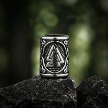 Silver Viking Valknut beard bead with intricate scrollwork, resting on dark rocks.