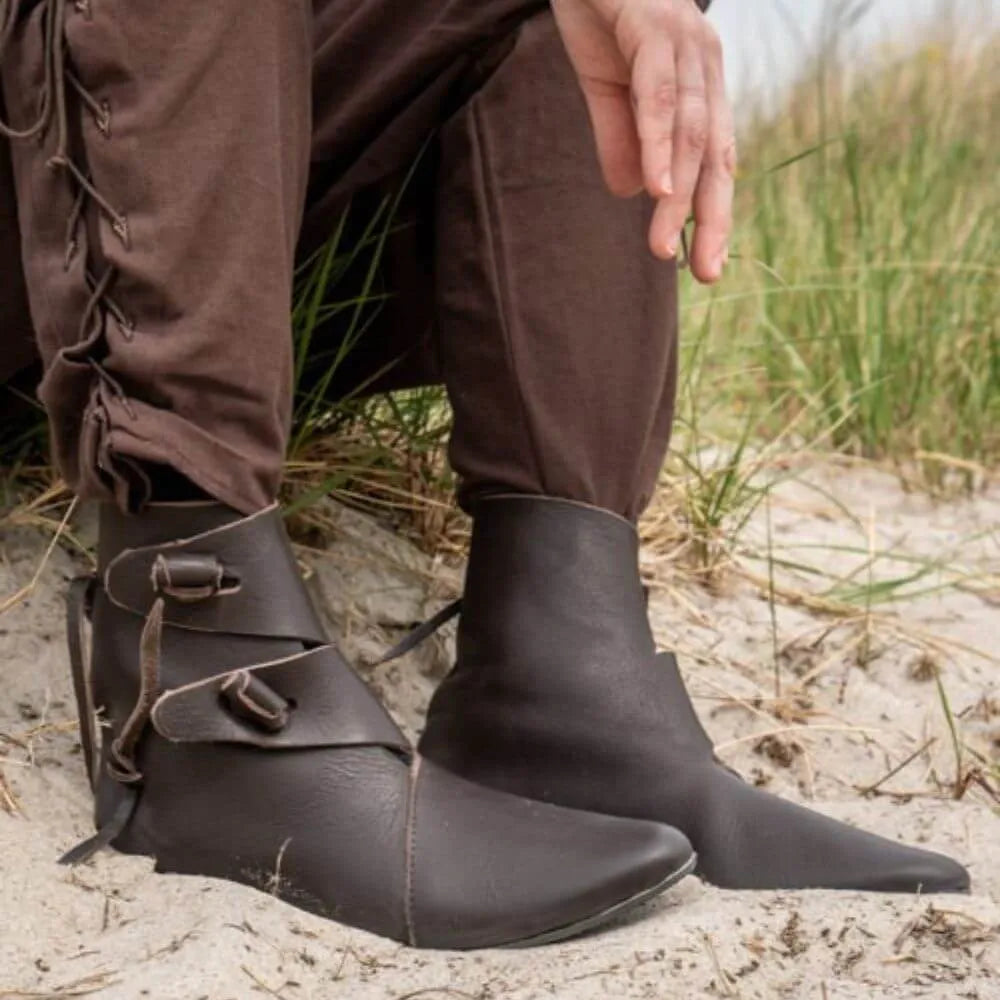 Brown leather Viking boots with buckle straps and pointed toes, worn with brown pants on sand.