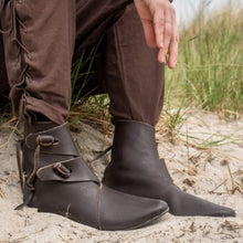 Brown leather Viking boots with buckle straps and pointed toes, worn with brown pants on sand.