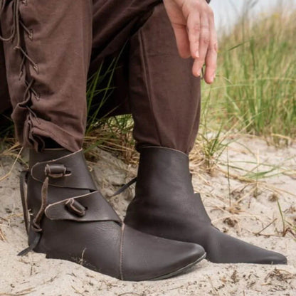 Brown leather Viking boots with buckle straps and pointed toes, worn with brown pants on sand.