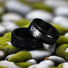 Two black Viking dragon rings, one with silver Celtic knot inlay, the other with subtle purple runes, on mossy rocks.