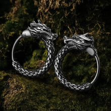 Pair of silver Viking dragon head hoop earrings with scale texture, resting on green moss.