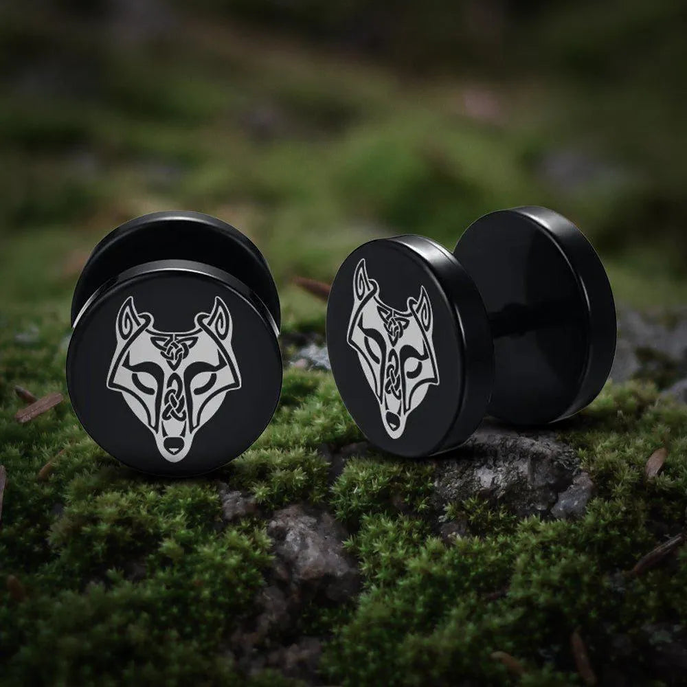 Black Viking stud earrings with a silver Fenrir wolf symbol, resting on mossy ground.