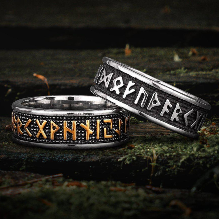 Two Viking Norse Futhark rings: one silver with black runes, one silver with gold runes, on mossy wood.