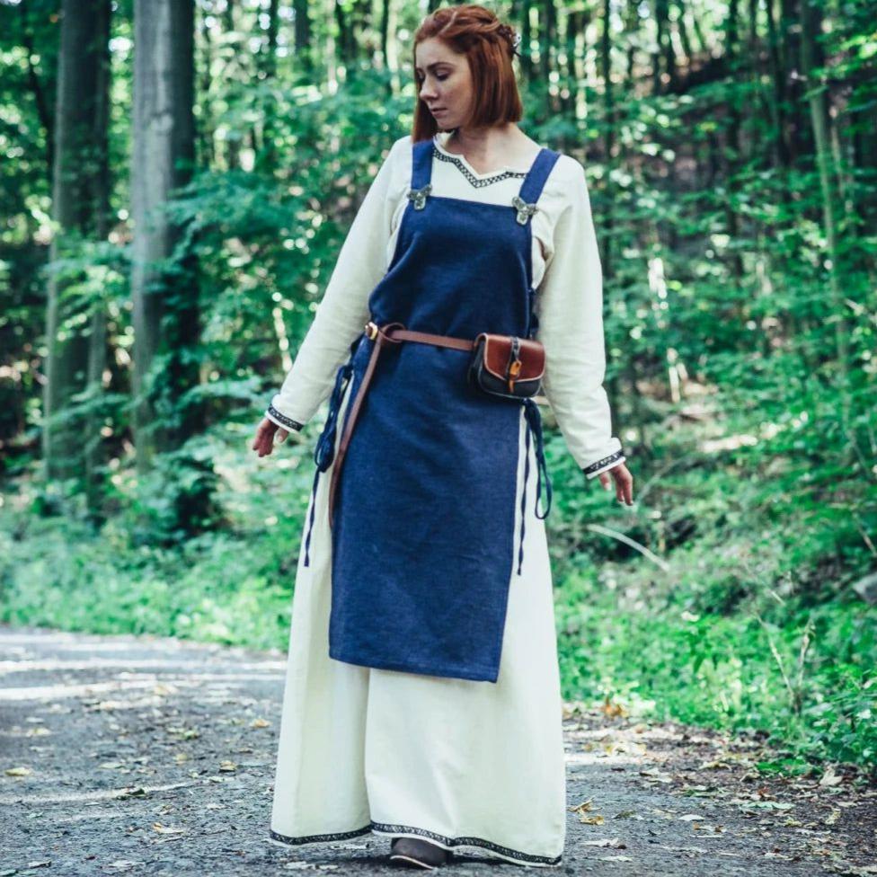 Viking woman in a cream cotton underdress and blue apron overdress with leather belt and pouch.