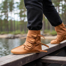Viking handmade tan leather boots with three buckles worn by a person walking on wood.