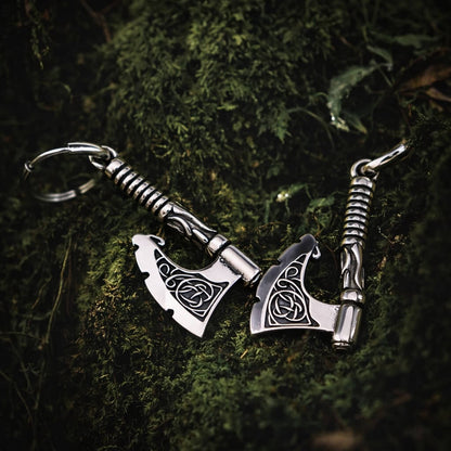 Viking Triquetra axe drop earrings, silver-toned metal, resting on green moss.
