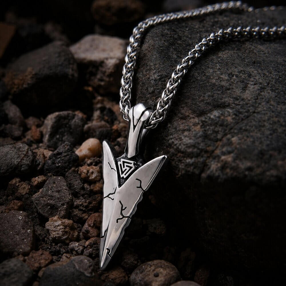 Viking Tyr arrowhead pendant necklace with Valknut design, silver-toned metal, on dark rocks.