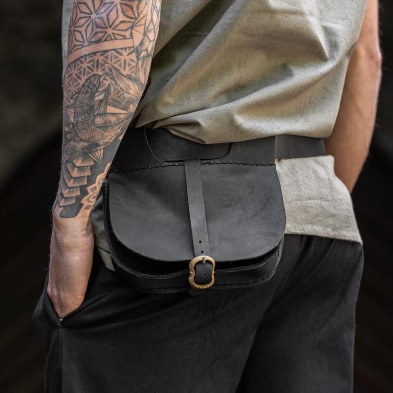 A Viking shown from the back wearing a black leather crossbody belt bag with a gold-tone buckle, styled with a beige sleeveless top and dark trousers, set against a blurred stone archway background.