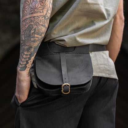 A Viking shown from the back wearing a black leather crossbody belt bag with a gold-tone buckle, styled with a beige sleeveless top and dark trousers, set against a blurred stone archway background.