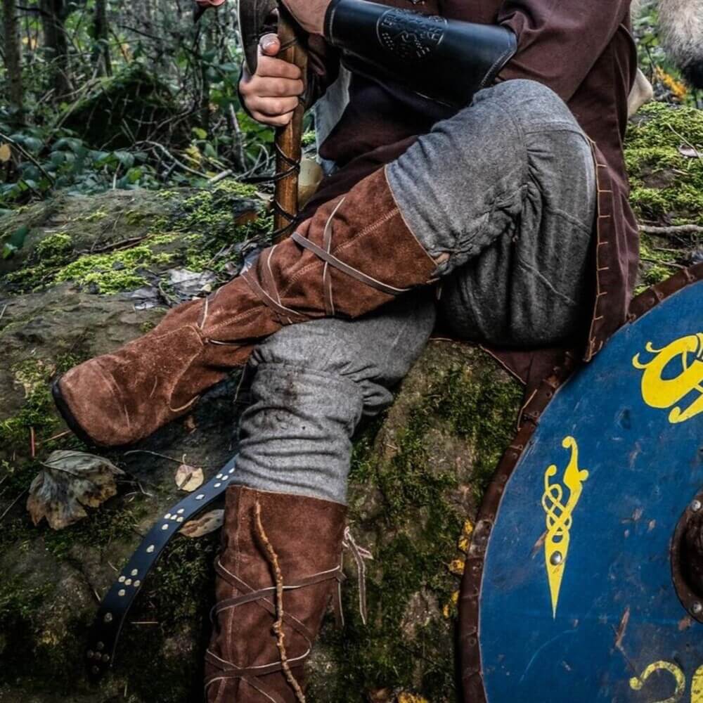 Person wearing brown suede Viking wrap-tied boots, seated on mossy rock beside a blue Norse shield in a forest.