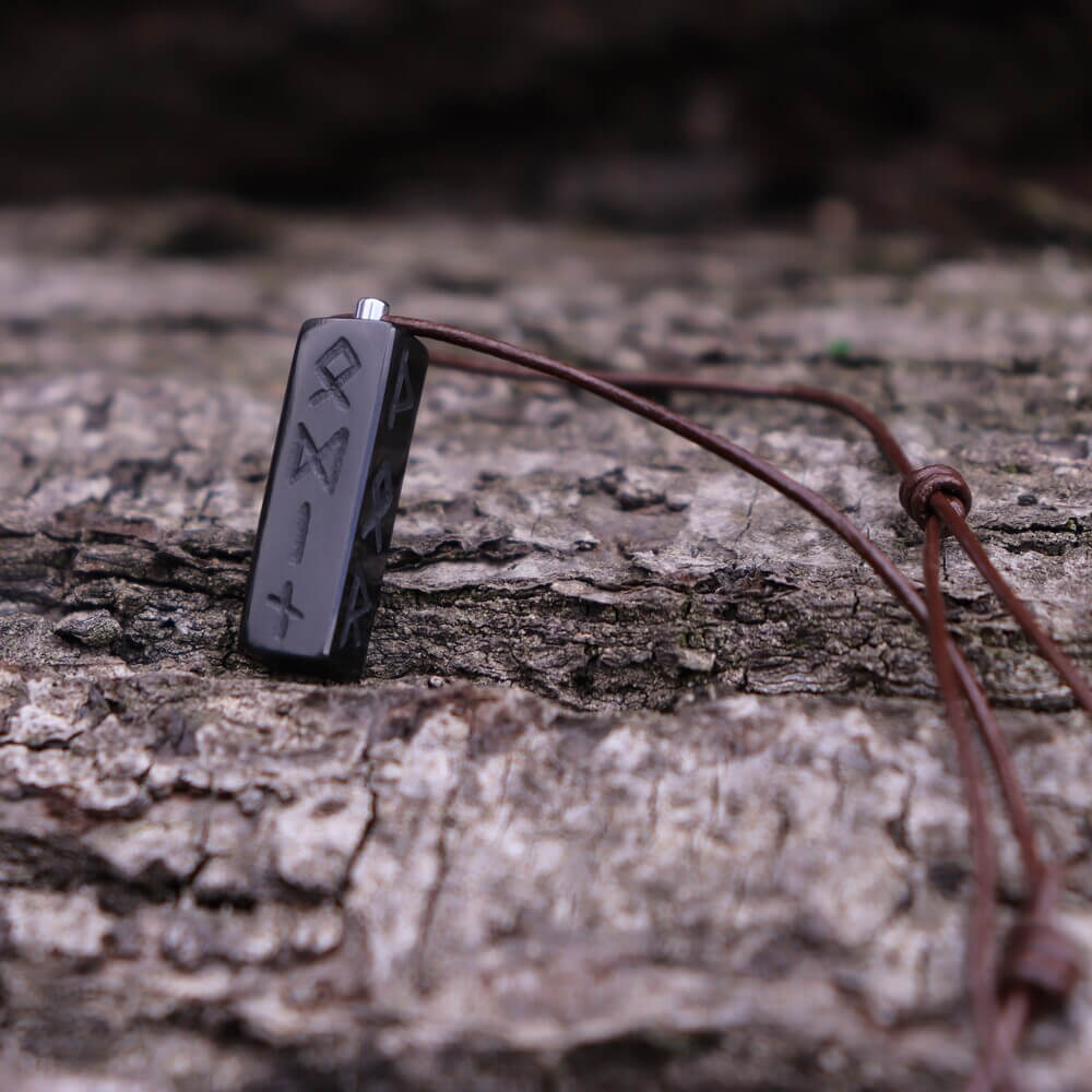 Runic Pillar Viking Necklace with carved runes on black pendant, hanging from brown cord over rugged tree bark.