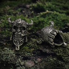 Two intricately carved Skull Viking Warrior beard beads rest on mossy ground, showcasing fierce Norse design and detail.