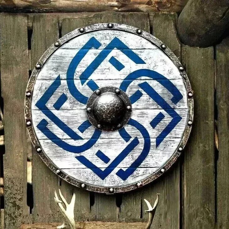 24-inch Viking medieval round shield featuring battle-worn distressed white painted wood with blue interwoven Celtic knot design – weathered steel rim with riveted studs and dark forged iron center boss – rustic wall décor mounted on aged wooden fence with deer antlers below