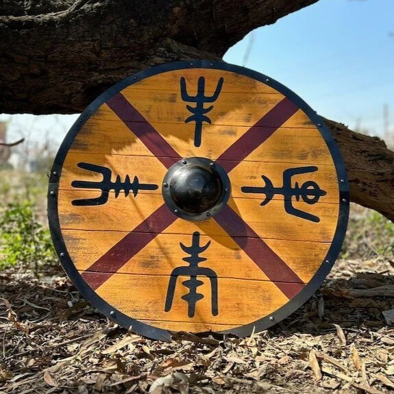 Viking round shield with golden birch wood, burgundy X-cross pattern, four Norse rune symbols, and black steel boss on outdoor wood chips